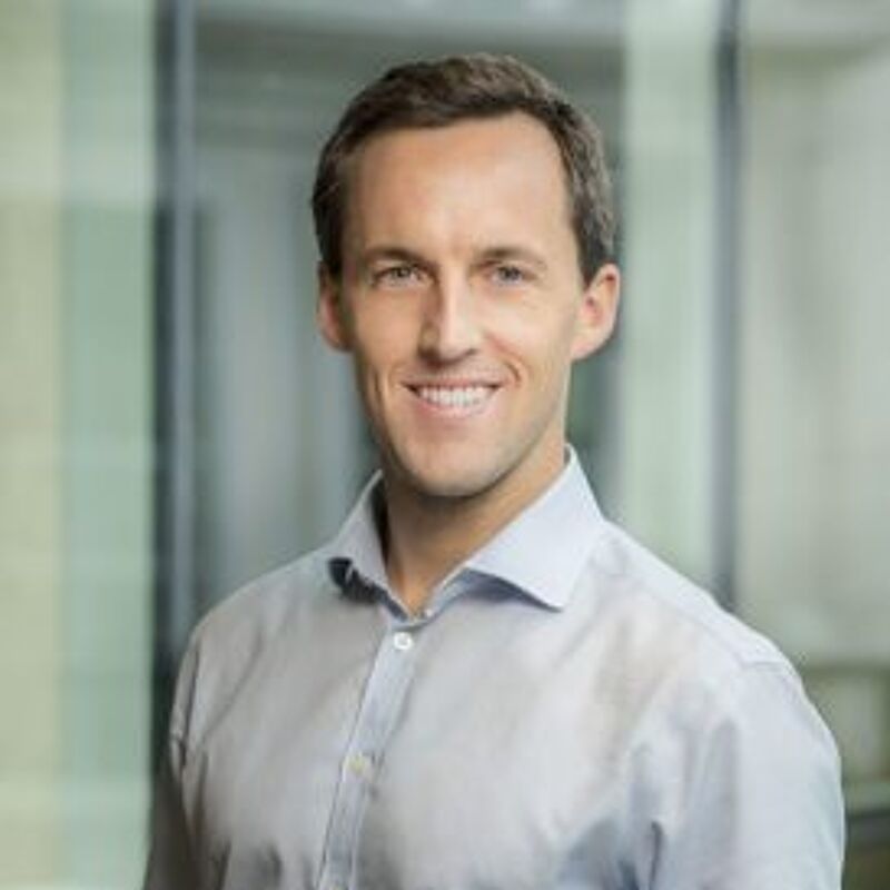 The image shows a man with short brown hair and a light blue shirt. He is smiling at the camera. The background is blurred, but it appears to be an office setting. The man looks to be in his late 30s or early 40s.